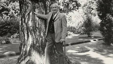 JRR Tolkien in the Oxford Botanic Garden with his right arm on the Black Pine in August 1973. The author is using a cane. The image is clak and white but it appears to have been a sunny day.