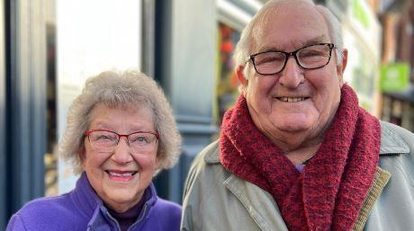 A woman wearing a purple jacket and red-rimmed glasses with curly grey hair. She is stood next to a man with a red scarf around his neck and a grey jacket. He has white hair and is wearing black rimmed-glasses. 