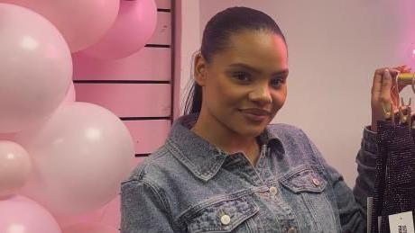 A woman with long black hair is standing between a pink balloon arch and a rack of sparkly jeans. She is wearing a denim jacket top and is wearing her hair slicked back.