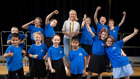 A group of 12 primary school children wearing blue t-shirts, stood on a stage with a woman in a white top holding a gold Olympic torch from the 2012 London Olympics. 