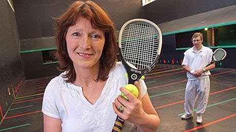 A woman with short red brown hair wearing a white t-shirt. She is standing on an indoor real tennis court which has colourful lines painted across the floor with numbers next to them. She is holding a small slim tennis-style racquet and a yellow tennis ball. There is a man standing in the background, also holding one of the racquets