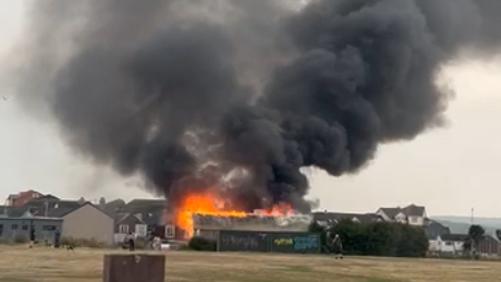 Large orange flames and thick smoke rise into the sky from the structure which is on fire. Buildings surround the fire site. A green grass area is at the front of the building.