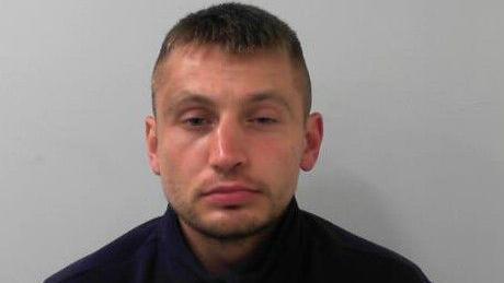 A police mugshot of a man in his twenties. He has short brown hair and his eyes are narrowed.