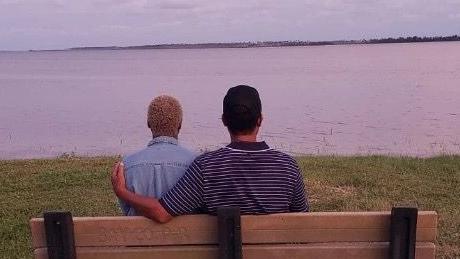 A man and a woman sat on a bench looking out to sea. The man has his arm around the woman's shoulders.