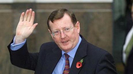 A middle aged man with brown hair and thin glasses waving at the camera. He has a navy suit on and blue shirt and maroon and blue striped tie. He is wearing a poppy.