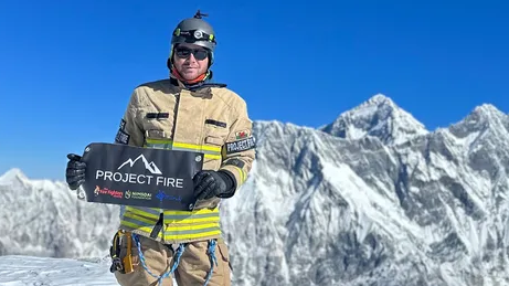 Rhys Fitzgerald in his firefighter gear standing on a mountain, with mountains and a clear blue sky behind him. He is holding a sign reading 'Project Fire'