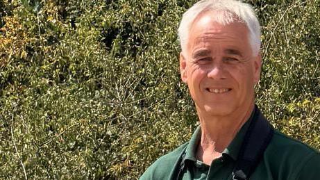 Dougal is standing on general grassland, with bushes behind him.
He is wearing a green t-shirt and a large watch on his left wrist.
