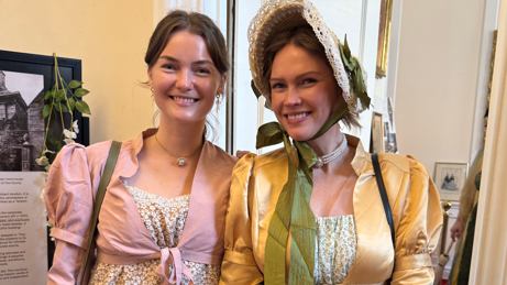 Two women wear floral dresses with cardigans over the top. Both are smiling and one wears a bonnet.