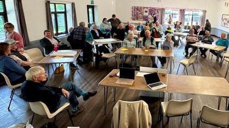 A room full of desks and chairs. There are people sat down and some stood. Some have laptops and notepads. The room has windows and white walls, it is sunny outside.