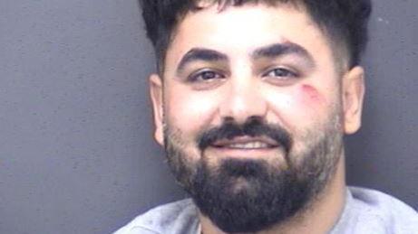 Police custody photo of Sarwar Rasool Mustafa. He is smiling and has a full black beard and hair and a slight graze on his left cheek.