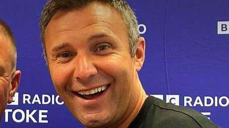 A man with short grey hair and wearing a black shirt smiles while standing in front of a purple banner that says "BBC Radio Stoke".