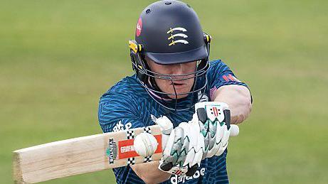 Ben Geddes hits a shot for Middlesex Cricket Club against Derbyshire in the 2025 One-Day Cup while wearing a dark blue top and black and yellow helmet