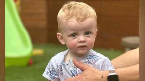 Oakley Barnett sitting on top of a large toy car in a garden. He is wearing a grey T-shirt and has blond hair.