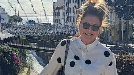 Elizabeth 'Liz' Goodall with long brown hair in a high pony tail wearing a white and black spotted blouse and sunglasses. She stands on a bridge above a stream with the sea in distance behind her on a sunny day. She is smiling.