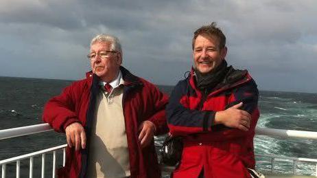 Two men standing on a ferry with the sea behind them