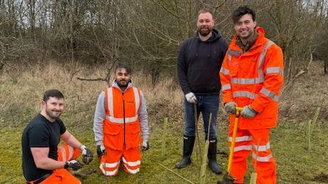 Four volunteers, looking at the camera, in an open part of a wooded area. A man on the left is kneeling down, wearing high-vis trousers and a black T-shirt, the next man is wearing orange high-vis trousers and gilet, the next man is wearing dark clothes and the man on the right is wearing high-vis orange trousers and a jacket. He is holding a spade. 