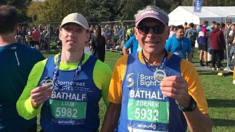 Liam Harkness and his guide Zdenek Gregor are pictured touching the medals that are hanging around their necks after completing a marathon together. They are wearing running vests. There are people gathered in a field behind them.