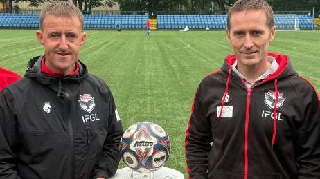 Vice-chairman Lee Dixon and Lawrence Looney standing either side of a football on a stand with the pitch at The Bowl stadium behind them. Both are wearing jackets with the FC Isle of Man logo on them.