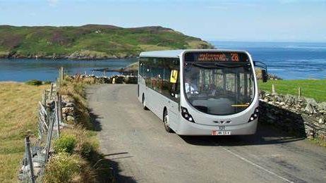 A silver, single-decker bus driving along a countryside road, with stone walls on either side and an island in the background. 