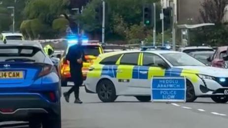 A police sign is in the middle of the road and says Heddlu. Police slow.  A police car is behind and a officer is pictured walking across the road