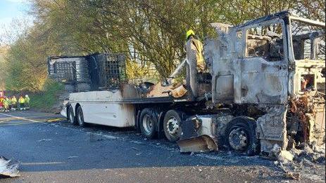 The aftermath of a lorry fire. The vehicle is large and has been completely burnt by a fire. It is parked in one lane of the M40 and firefighters can be seen in attendance.