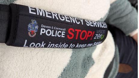 A black cover is attached to a seat belt in this close up shot. The words Emergency Services, Devon and Cornwall Police and Look inside to keep me safe are written on the cover. in white on a black background.
The word Stop is written in red.