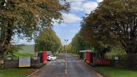 The entrance to the business can be seen with a red gate open and a red sign for Ricoh on the right hand side. A long straight road is lined with three on either side and a car is parked on the left side just inside the entrance.