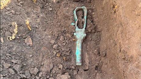 A green metal rattle can be seen in an archaeological trench.