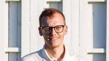 Luke Terry stands in front of a cricket club pavilion. He wears cricket whites and glasses. He has short, light brown hair.