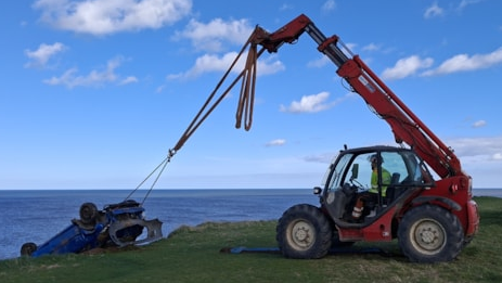 An upturned blue Peugeot being hauled to safety from a cliff by a red tractor. A recovery lorry is parked behind the tractor. White clouds are dotted in the blue sky. The blue sea can also be seen from over the cliff. 