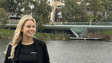 A woman wearing a black top and T-shirt ,stands alongside a body of water. There is a bridge and a building in the background. She has long blonde hair, and is smiling as she looks to the right