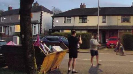 Door cam footage of the two men responsible for fly-tipping. They both were dark shorts, one has a white T-shirt the other has a black T-shirt. They a re standing next to a yellow skip on a suburban street