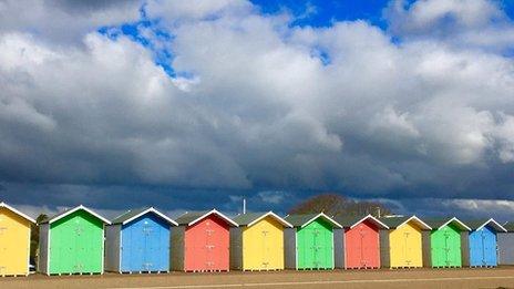 A row of yellow, green, blue and red beach huts sit along the front with big white clouds above and blue sky at the top