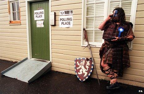 A man dressed as William Wallace leans against a voting booth on his mobile phone