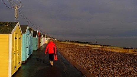 A person walks past coloured beach huts on the left with the beach and sea to the right with grey skies above