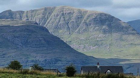 A blue/grey sky forms the back drop to a Scottish mountain range with a cottage in the foreground.