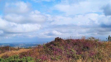 The top of a hill with fields in the far background and blue skies and clouds overhead.