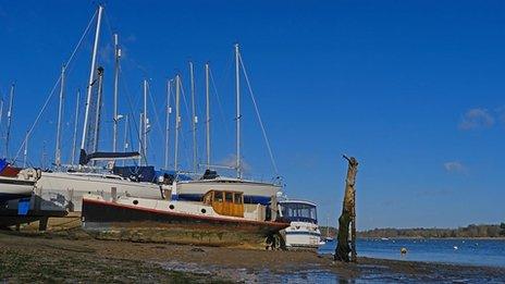 Sail boats sit moored on the shore with a bright blue sky over head and blue sea to the right