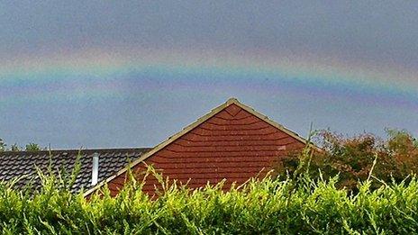 A rainbow sits in a blue sky across the roof of a house