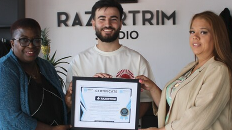 Razwan stands in between two ladies and the three of them hold the framed certificate he was given in his salon. One woamn on the left is wearing a navy outfit, Razwan has a white t-shirt on, and has dark hair and a beard and the woman on the right is wearing beige clothing.
