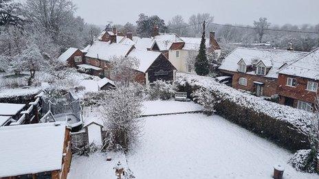 A garden stretches out covered in snow with a building to the left, a long hedge to the right with houses behind it and to the rear. The horizon is covered in trees and the sky is grey behind