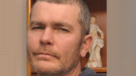 Abraham Cilliers stands in a room beside a shelf with a figurine. He has a neutral expression and has short grey-brown hair and stubble.