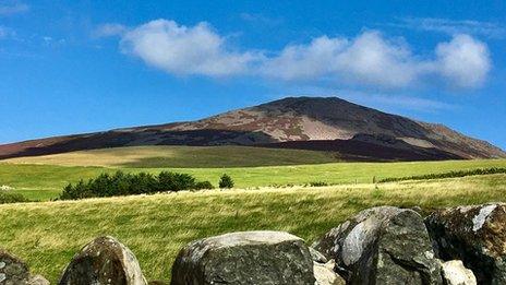 A mountain with green fields and a stone wall in front and bright blue sky above.