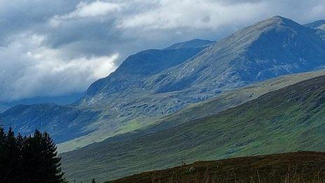 Grey and white clouds in the sky above blue and green mountains wit a field in the foreground.