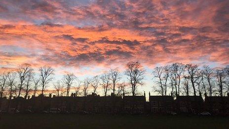 Clouds turn red and blue as the sun sets over a row of trees and houses