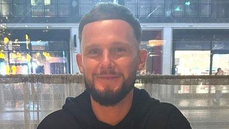 Daniel Conway wearing a black hoodie and smiling at the camera. He has dark brown hair and a trimmed beard, and is sitting in front of a gauze curtain at a bar.