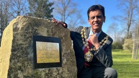 The Mayor of Oldham Dr Zahid Chauhan rests on his haunches next to the memorial boulder. He is wearing his mayoral chain. The boulder has in inscription on it that cannot be made out in the photograph. 