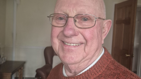 Bald-headed Bob Leigh, wearing glasses and a reddish brown jumper, smiles for the camera at home