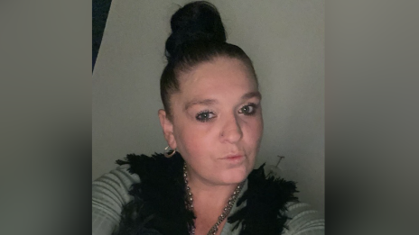 A woman with dark hair in a bun wears grey and black and looks at the camera taking a selfie.