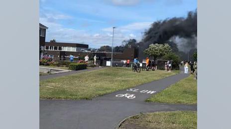 A picture of Shanklea Primary School during the fire. Black smoke is billowing from the building and blowing to the right. A small group of people has gathered outside the school to watch.